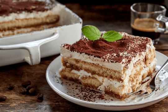 Italian Tiramisu on a white plate placed on a dark brown table and a black coffee in the back.